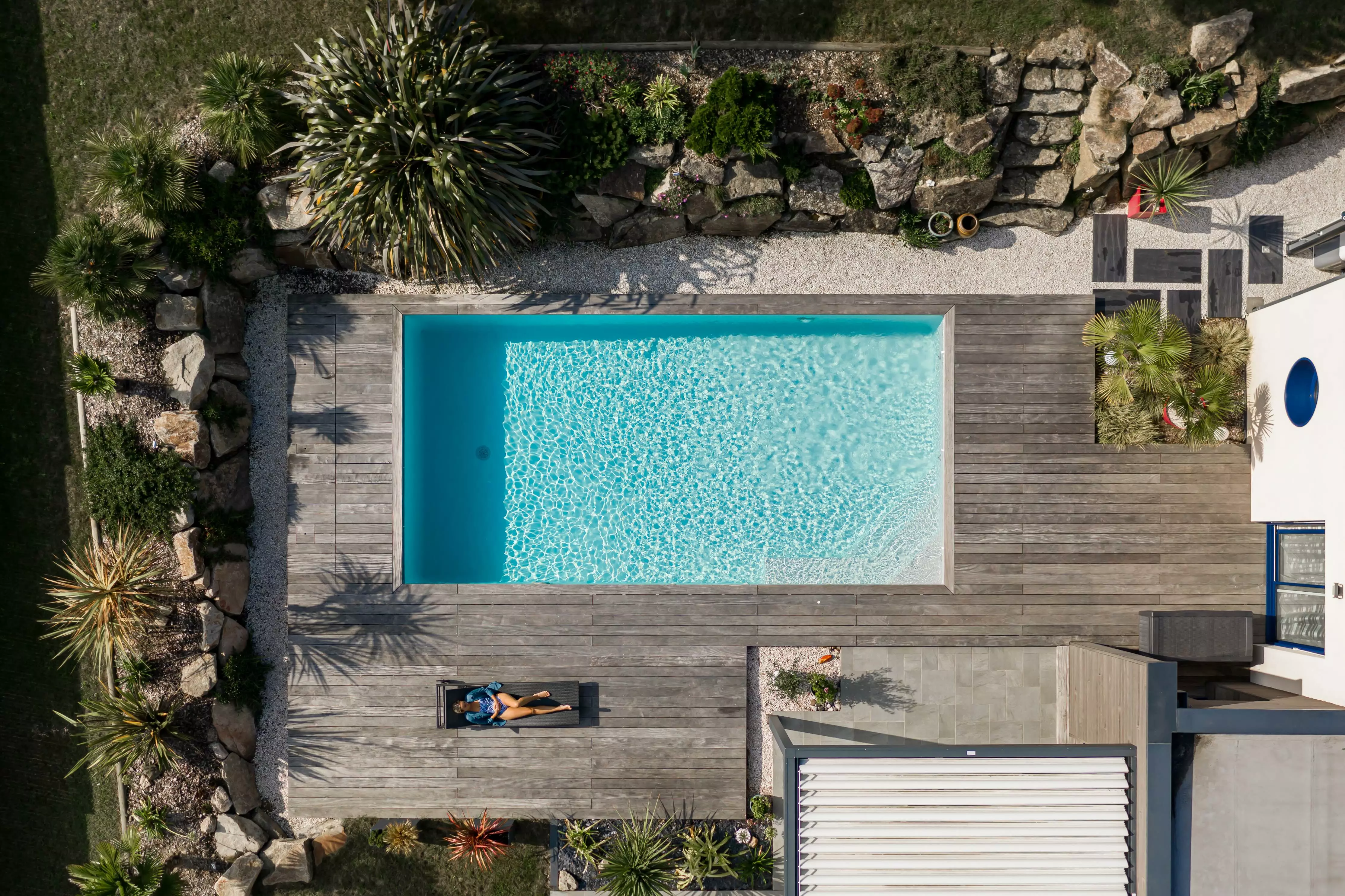 Une habitation où la piscine est au cœur, connectée à un espace rocheux par une terrasse, le tout décoré de plantes exotiques.