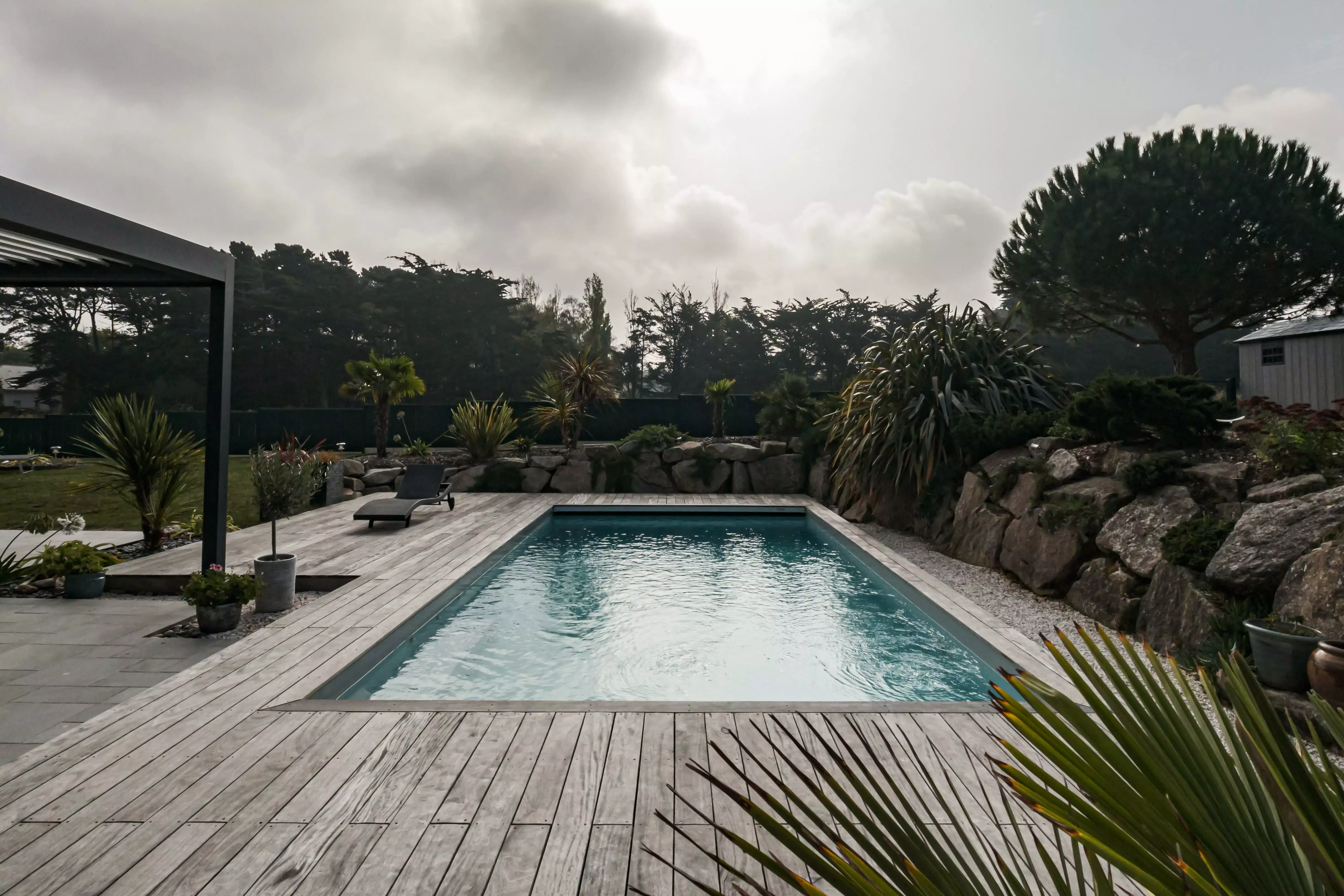 Une maison contemporaine dessinée avec la piscine en focus, adossée à un espace rocheux via une terrasse, rehaussée par des plantes venues d'ailleurs.